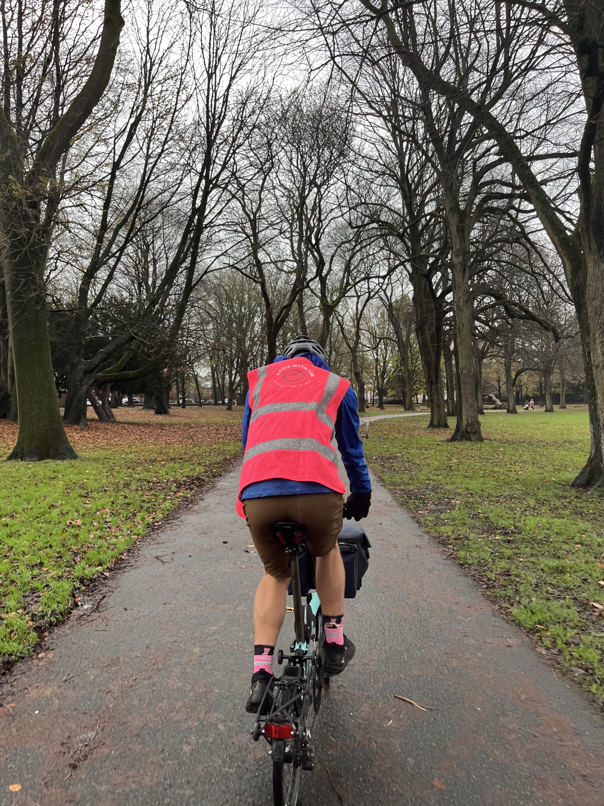 An adult cycling away from the camera in a park wearing a pink hi viz vest
